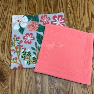 May include: Two fleece pet bed pads. One is coral pink with a stitched border. The other is gray with a floral pattern of white, pink, and red flowers, green leaves, and red berries. The text "teachersPiggyPet Shop" is visible.
