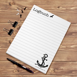 Op de afbeelding: Een wit, gestreept notitieboekje met het woord "Logbuch" bovenaan en een zwart anker met touw onderaan. Het notitieboekje heeft een zwart vogelsilhouet in de rechterbovenhoek. De tekst "www.grafikundmehr.de" staat onderaan het notitieboekje.