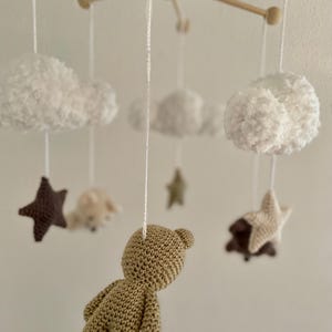 Könnte beinhalten: Handgefertigtes Baby-Mobile mit Holzrahmen und gehäkelten Elementen. Es zeigt weiße Wolken, braune und beige Sterne sowie einen beigen Teddybär. Die sanften, neutralen Farben schaffen eine beruhigende Dekoration für das Kinderzimmer.