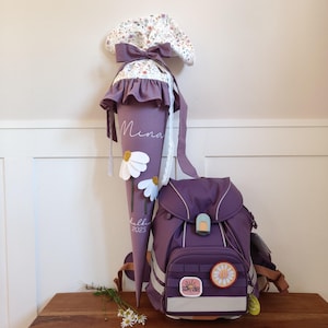 Könnte beinhalten: Ein lila Schultüten- und Rucksack-Set. Die Schultüte hat ein blumiges Muster auf der Oberseite, eine große Schleife und den Namen "Mina". Der Rucksack hat einen Gänseblümchen-Aufnäher und die Worte "Happy Vibes".