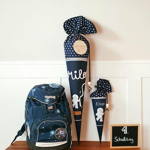 Könnte beinhalten: Ein marineblauer Rucksack und zwei Schultüten mit Weltraum-Motiv. Die Tüten sind mit weißen Sternen und Astronauten-Illustrationen verziert. Eine Tüte trägt den Namen "Milo" und die andere "Elias". Eine kleine Tafel zeigt "1. Schultag".