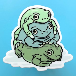 May include: A sticker featuring three green frogs stacked on top of each other. The top frog is green with black eyes and a wide smile. The middle frog is blue with black eyes and a closed mouth. The bottom frog is green with black eyes and a closed mouth.