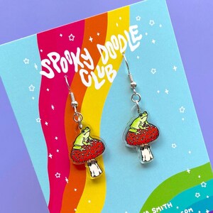 May include: A pair of silver earrings with a green frog sitting on a red mushroom. The earrings are displayed on a rainbow background with the text "Spooky Doodle Club" and "By Elaina Smith Spookydoodleclub.etsy.com".
