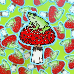 Pode incluir: Um sapo verde sentado em um cogumelo vermelho e branco com manchas brancas. O cogumelo tem um caule branco com uma franja na parte inferior. O fundo é um padrão de cogumelos vermelhos e brancos.