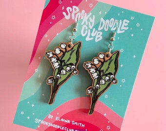 May include: A pair of wooden earrings featuring a lily of the valley design. The earrings are painted with green leaves and white flowers. The earrings are on a card that says "Spooky Doodle Club" and "By Elaina Smith Spookydoodleclub.etsy.com".
