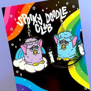 May include: Earrings from Spooky Doodle Club, featuring two cartoon Furby-like characters. One is purple with angel wings and a halo, the other is blue with candles. The background is black with a rainbow and stars.
