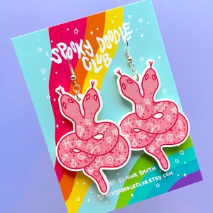 May include: Pink acrylic earrings shaped like two-headed snakes with white astrological symbols and a silver hook. The earrings are displayed on a card with the text "Spooky Doodle Club" and "By Elaina Smith"