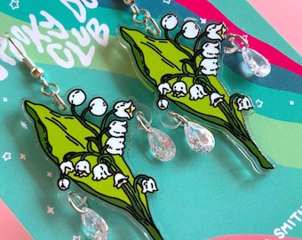 Op de afbeelding: Een paar hangende oorbellen met doorzichtige acryl bedels in de vorm van lelietjes-van-dalen met groene bladeren en witte klokvormige bloemen. Elke oorbel bevat een kleine, heldere, traanvormige kristallen bedel bevestigd aan een zilveren metalen ring.