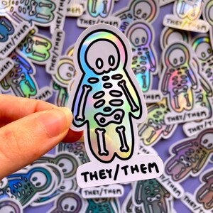 May include: A holographic sticker with a skeleton figure and the text "THEY/THEM". The sticker is surrounded by other similar stickers with different designs.
