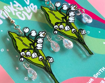 May include: A pair of dangle earrings featuring a cluster of white lily of the valley flowers with green leaves and clear crystal teardrop accents. The earrings are made of acrylic and have silver hooks.