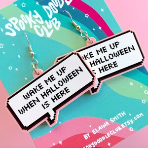 May include: Pink and white statement earrings with a pixelated speech bubble design. The text on the earrings reads "Wake me up when Halloween is here".