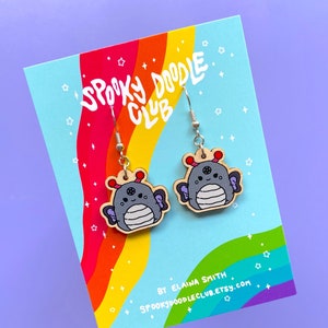 May include: A pair of wooden earrings featuring a cute cartoon creature with wings. The earrings are painted in shades of grey, white, and purple. The card reads "Spooky Doodle Club" and "By Elaina Smith Spookydoodleclub.etsy.com".