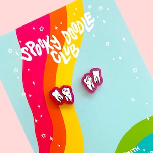 May include: A pair of pink tooth earrings with white enamel detailing. The earrings are displayed on a card with a rainbow background and the text "Spooky Doodle Club" and "By Elaina Smith Spookydoodleclub.etsy.com".