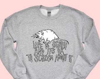 Könnte beinhalten: Ein graues Sweatshirt mit einer schwarz-weißen Illustration eines schreienden Opossums. Der Text "Life is scary and it's ok to scream about it" ist unter dem Opossum gedruckt.