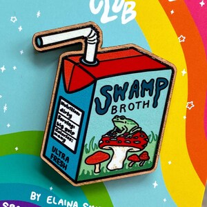 May include: A wooden pin shaped like a carton of swamp broth with a green frog sitting on a mushroom. The carton has a straw sticking out of it and says "Swamp Broth" on the front. The pin is on a card with a rainbow background and the text "Spooky Doodle Club" at the top.