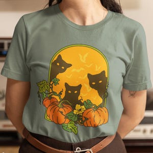 以下が含まれることがあります： ハロウィーンのデザインが施されたセージグリーンのTシャツ。グラフィックには、黒い猫のシルエット、オレンジ色のカボチャ、コウモリのいる黄色の月が含まれています。「SPOOKY DOODLE CLUB」の文字が左下にプリントされています。