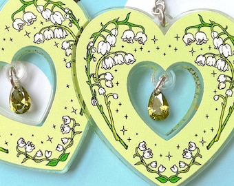 May include: A pair of green heart-shaped earrings with a lily of the valley design and a green crystal charm. The earrings are on a white background with the text "Spooky Doodle Club".