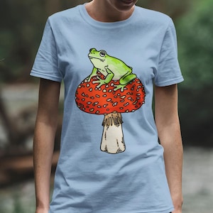 May include: A light blue t-shirt with a green frog sitting on a red and white mushroom.