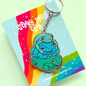 May include: A wooden keychain featuring three stacked frogs in shades of green and blue. The keychain is attached to a silver key ring and has the text "Spooky Doodle Club" printed on the card it is attached to.
