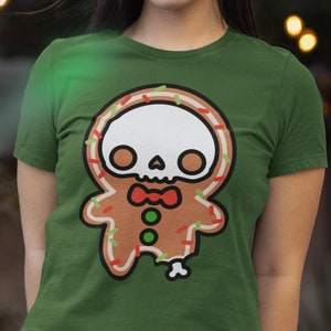 May include: Green t-shirt featuring a gingerbread man design with a skull face, a red bow tie, and colorful sprinkles. The gingerbread man has a missing leg. The shirt is from Spooky Doodle Club.