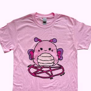 May include: Pink t-shirt featuring a cartoon illustration of a purple and pink creature with wings, sitting on a pink pentagram.