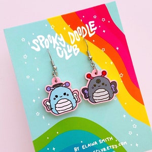 May include: Dangle earrings with cartoon creatures in blue and grey, pink accents, and silver hooks. The earrings are presented on a card with the text "Spooky Doodle Club" and a rainbow design.
