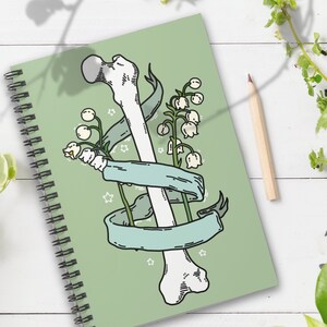 Op de afbeelding: Een groen spiraalnotitieblok met een illustratie van een wit bot en lelietjes-van-dalen bloemen. Het bot is omwikkeld met een blauw lint en er zijn witte sterren op de achtergrond.