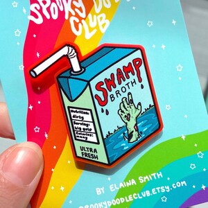 May include: A colorful enamel pin featuring a cartoon illustration of a green and blue juice box with a red straw. The juice box has the words "Swamp Broth" printed on it and a zombie hand sticking out of the liquid. The pin is on a card with the words "Spooky Doodle Club" and "By Elaina Smith" printed on it.