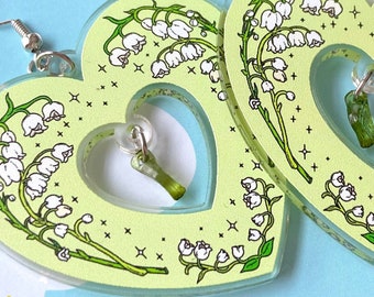 May include: A pair of green heart-shaped earrings with a lily of the valley design and a clear crystal charm. The earrings are on a white background with the text "Spooky Doodle Club".
