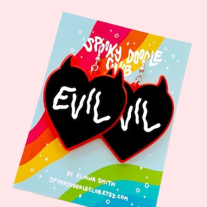 May include: A pair of black heart-shaped earrings with red outlines and silver hooks. The earrings have the word "EVIL" written in white on each heart. The earrings are displayed on a blue and rainbow background with the text "Spooky Doodle Club" and "By Elaina Smith Spookydoodleclub.etsy.com".