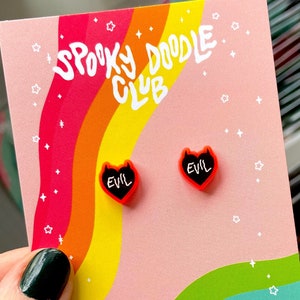 May include: A pair of black and red heart-shaped earrings with the word "EVIL" written in white on each heart. The earrings are displayed on a pink and rainbow background with the text "Spooky Doodle Club" and "By Elaina Smith" written in white.