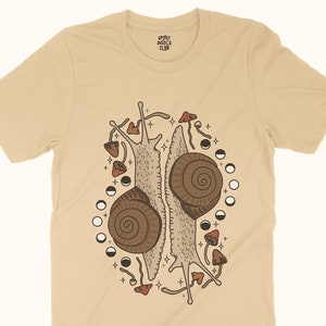 Pu&ograve; includere: Una maglietta beige con un design grafico di due lumache con i loro gusci uno di fronte all'altro. Le lumache sono circondate da funghi e lune crescenti. Il testo "Sleepy Doodle Club" &egrave; stampato sulla maglietta.