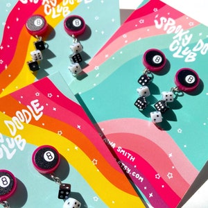 Op de afbeelding: Een paar kleurrijke oorbellen met een roze 8-ball charme en een hangende ketting van zwarte en witte dobbelstenen. De oorbellen worden getoond op een kaart met de tekst "Spooky Doodle Club".