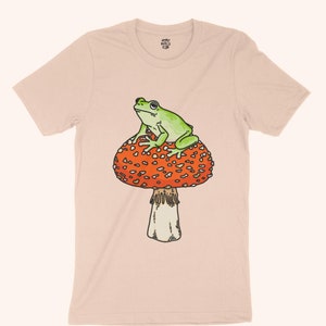 May include: A light pink t-shirt with a green frog sitting on top of a red and white mushroom.