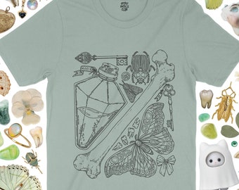 May include: A sage green short-sleeve t-shirt featuring a black line art graphic of a potion bottle, a large bone, a beetle, a moth, two ornate keys, a small bow, and several teeth. The inner neck label displays the text Spooky Doodle Club.