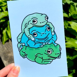 May include: A blue background with a stack of three cartoon frogs. The top frog is green, the middle frog is blue, and the bottom frog is green. The frogs are all smiling.