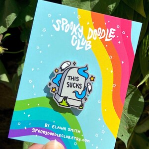 May include: A blue and white enamel pin featuring a tombstone with the text "This Sucks" and ghosts. The pin is on a rainbow background with the text "Spooky Doodle Club" and "By Elaina Smith" in black.