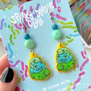 May include: A pair of dangle earrings featuring a stack of three cartoon frogs in shades of green and blue on a yellow base. The earrings have a light blue and green bead accent and are attached to a silver hook. The background is a light blue card with the text "Spooky Doodle Club".