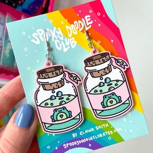 May include: A pair of pink and green frog in a jar earrings. The earrings are made of acrylic and feature a cute frog inside a glass jar with bubbles. The earrings are on a white card with the text "Spooky Doodle Club" and "By Elaina Smith" printed on it.