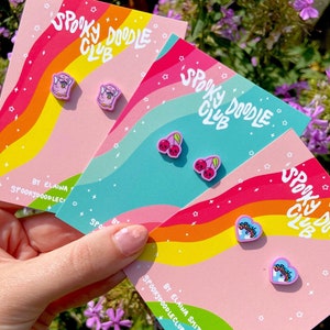 May include: Three cards with a rainbow background and stars. Each card features a pair of pink and purple earrings with different designs. The earrings are shaped like a ghost, a cherry, and a heart. The text on the cards reads "Spooky Doodle Club" and "By Elaina Smith, SpookyDoodleClub.etsy.com".