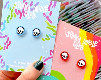 May include: Two pairs of skull stud earrings on branded display cards. One pair features white skulls with black outlines on light blue bases, while the other pair has white skulls with black outlines on bright red bases. Cards read Spooky Doodle Club.