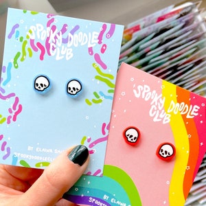 May include: Two pairs of skull stud earrings on branded display cards. One pair features white skulls with black outlines on light blue bases, while the other pair has white skulls with black outlines on bright red bases. Cards read Spooky Doodle Club.