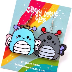 May include: A pair of acrylic dangle earrings depicting cartoon alien-like creatures. One is light blue with a halo, the other is grey with a pentagram. They hang from silver hooks on a card with a rainbow design and the text "Spooky Doodle Club".
