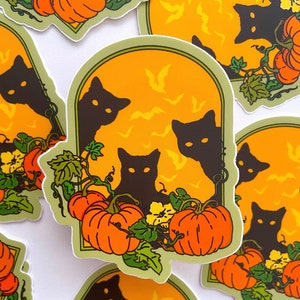以下が含まれることがあります： オレンジ色のカボチャと黄色の花が絡まる蔦の後ろから、黒い猫が3匹顔をのぞかせています。背景は明るいオレンジ色で、コウモリが空を飛んでいる模様が描かれています。画像は緑色のアーチで囲まれています。