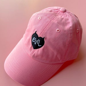 May include: A pink baseball cap with a black heart embroidered on the front. The heart has the word "EVIL" written in white inside.