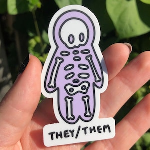 May include: A purple skeleton sticker with a skull head and the text "THEY/THEM" on a white background.