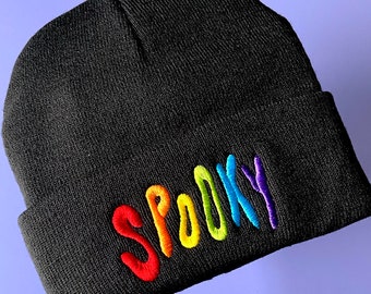 Pode incluir: Um gorro de malha preto com a palavra SPOOKY bordada num gradiente de arco-íris vibrante com fios vermelho, laranja, amarelo, verde, azul e roxo. O texto está posicionado na dobra frontal do gorro, que tem um acabamento de tecido texturizado.