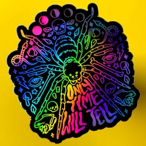 May include: A holographic sticker with a rainbow spider design and the text "Only time will tell". The spider is surrounded by various symbols, including skulls, eyes, and stars.
