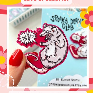 May include: A pink and white cartoon possum with a speech bubble that says "#%$!?"  The possum is holding its paws up in a defensive position. The possum is on a pink background with a white outline. The text "Spooky Doodle Club" is visible in the background.