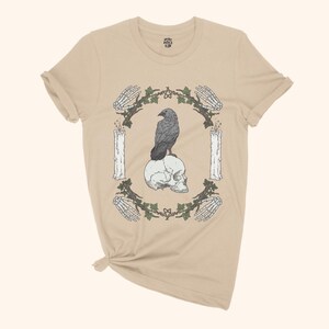 May include: Beige t-shirt featuring a graphic design with a raven perched on a skull, surrounded by candles, skeleton hands, and a leafy wreath. The shirt has short sleeves and a crew neck.